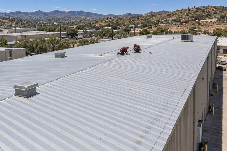 Two roofers replacing screws along the ribs of a metal warehouse roof