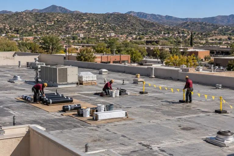 Three roofers staging membrane rolls behind a flagged warning line on a flat roof